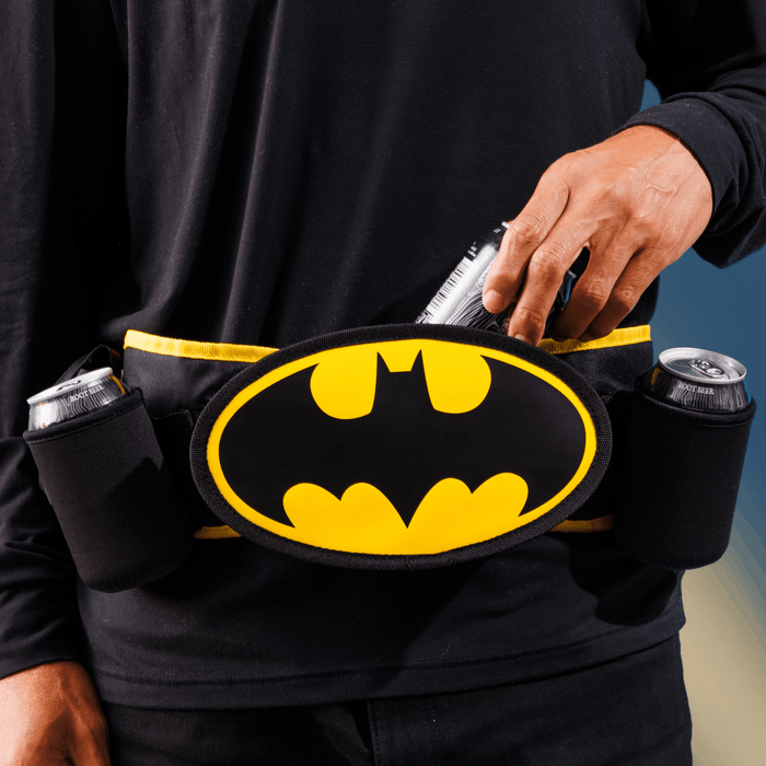 A close-up of a person wearing a black cooler belt with a large yellow Batman logo in the center and black can holders on both sides, with the person's hand reaching into the center cooler compartment.