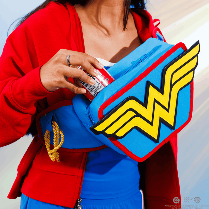 A close-up of a person wearing a red and blue superhero-style outfit and holding a matching blue cooler belt with a large Wonder Woman logo. The person is inserting a drink into the pouch.