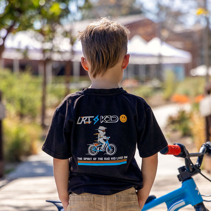 A young boy wearing the LRT x SUPER73 Limited Edition Collab Tee facing away from the camera. The LRT x K1D logo is visible on the back of the tee and a Blu Tang K1D ebike is parked in front of the boy.
