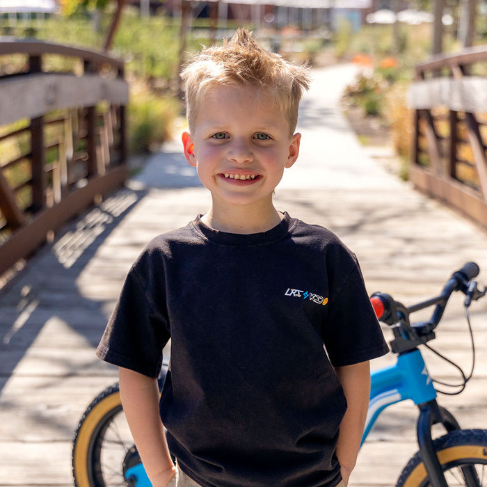 A young boy wearing the LRT x SUPER73 Limited Edition Collab Tee facing the camera. The LRT x K1D logo is visible on the left chest area of the tee and a Blu Tang K1D ebike is parked behind the boy.