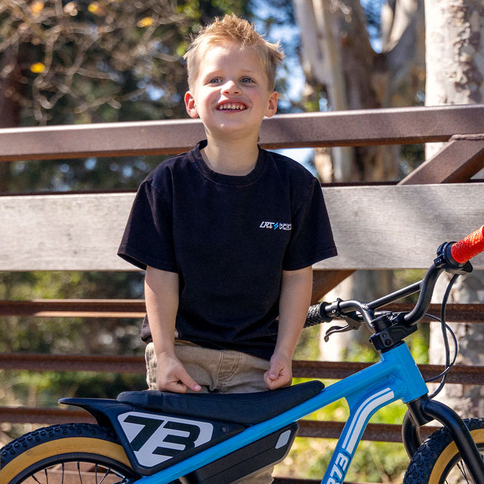 A young boy wearing the LRT x SUPER73 Limited Edition Collab Tee facing the camera and smiling. The LRT x K1D logo is visible on the left chest area of the tee and a Blu Tang K1D ebike is parked in front of the boy as he stands in front of a fence.