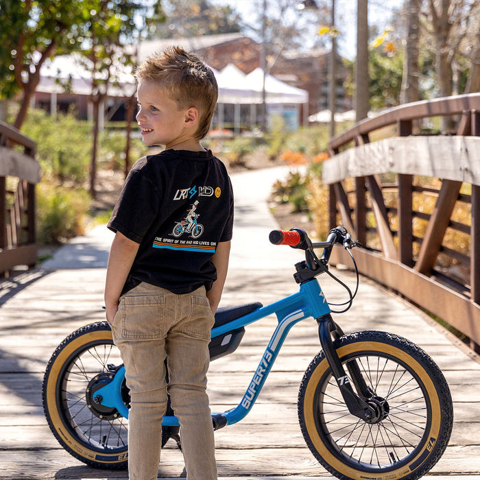 A young boy wearing the LRT x SUPER73 Limited Edition Collab Tee with his head turned to the left. The LRT x K1D logo is visible on the back of the tee and a Blu Tang K1D ebike is parked in front of the boy.
