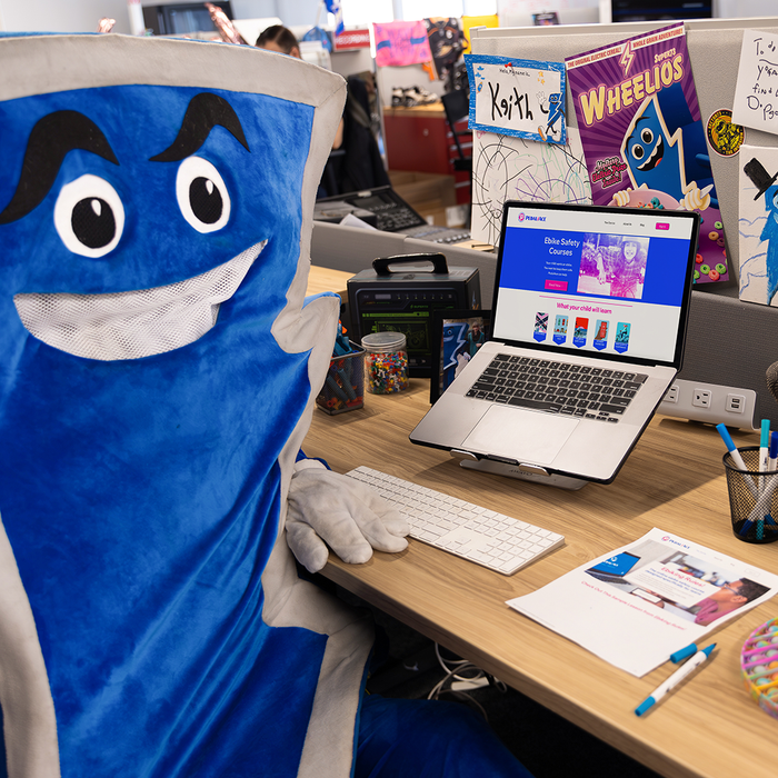 Keith, the SUPER73 mascot, sitting at a desk with a laptop displaying the PedalAce website and surrounded by various office supplies and documents.