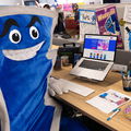 Keith, the SUPER73 mascot, sitting at a desk with a laptop displaying the PedalAce website and surrounded by various office supplies and documents.