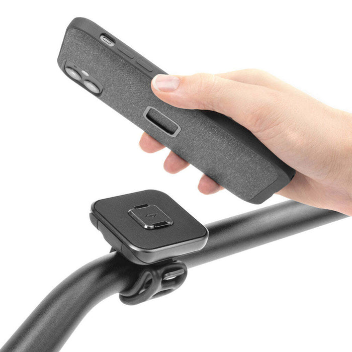 Close-up of the Universal Bar Mount installed on a bike handlebar. A hand is holding an iPhone to demonstrate where the mount would connect to the back of the iPhone case.